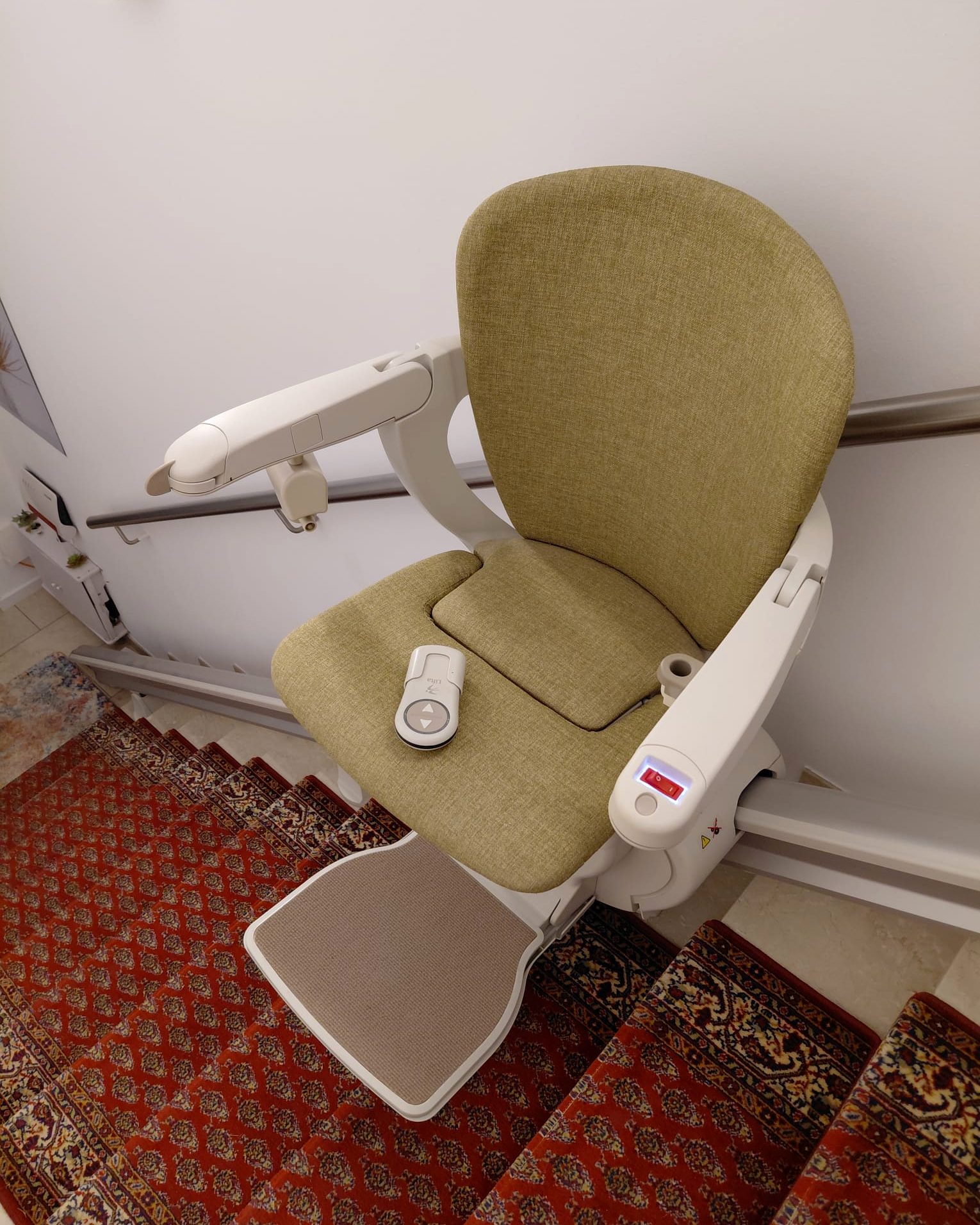 Eingebauter Treppenlift mit hellgrünem Sitz und Fernbedienung auf einer Treppe mit rotem Läufer